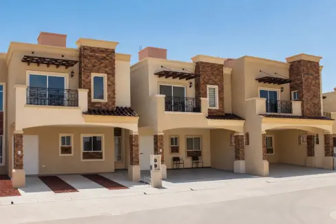 Row of modern beige townhouses with stone columns and black metal balconies against a clear blue sky.