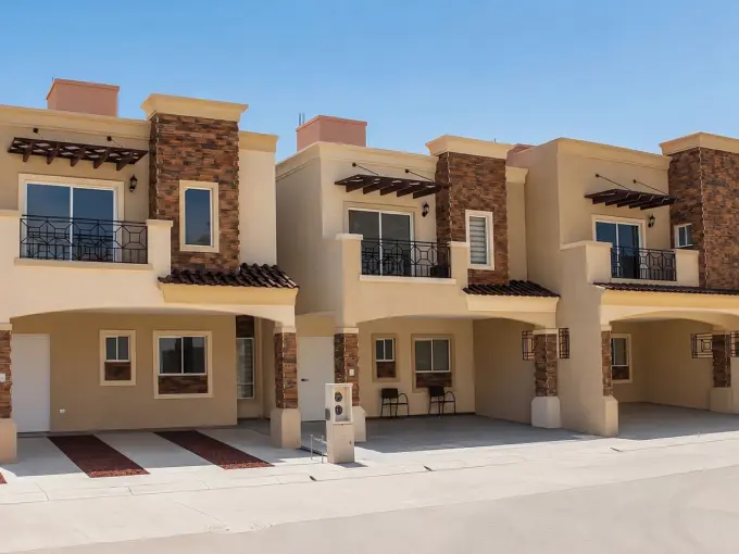 Row of modern beige townhouses with stone columns and black metal balconies against a clear blue sky.